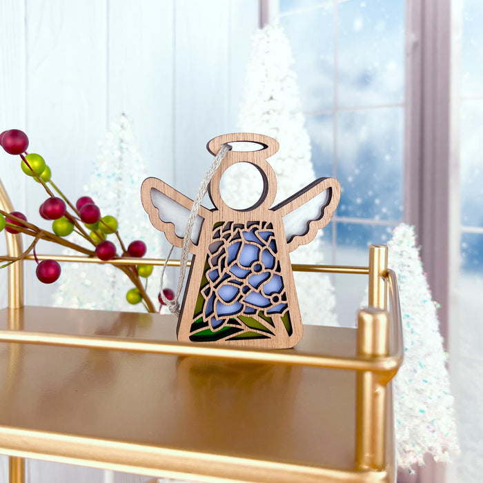 A delicate wooden angel ornament from Forged Flare®, known as the Hydrangea Ornament | 3.5" Angel Figurine | Mother's Angels®, perfect for your angel figurines collection, with wings, a halo, and a cutout pattern filled with purple and green stained glass–style details, rests on a gold shelf. In the background, snowy trees, red berries, and frosted window panes contribute to a festive atmosphere.