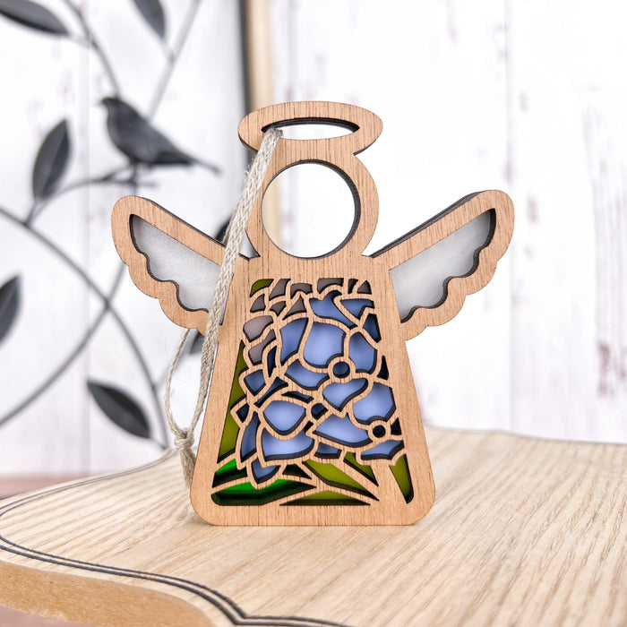 A 3.5" wooden angel figurine with intricate floral cutouts, part of the Mother's Angels® collection from Forged Flare®, reminiscent of the Hydrangea Ornament, stands on a light wooden surface. The floral design in the angel's dress is highlighted with blue and green stained glass–style elements. In the blurred background, a metal branch with a bird silhouette is visible.