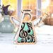 A 3.5" Music Ornament Angel Figurine from the Mother's Angels® collection by Forged Flare®, adorned with intricate designs and musical notes, hangs from a braided linen string against a light background. Behind it, large windows showcase an autumn scene with blurred pumpkins and vases, enhancing the cozy atmosphere—making it an ideal gift for music teachers.