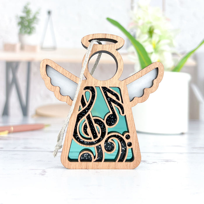 A small Music Ornament from the Mother's Angels® collection by Forged Flare®, featuring intricate patterns and teal accents, sits on a white surface. In the background, there's a blurred white pot with green leaves and white flowers, accompanied by some wooden and metal objects, creating a rustic ambiance—an ideal gift for music teachers or a charming addition to your collection of angel figurines.