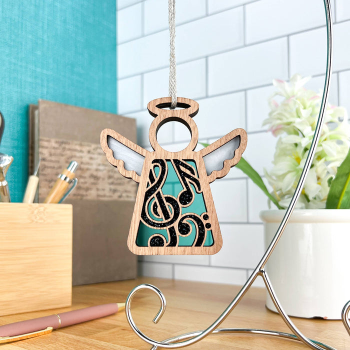 A Forged Flare® Music Ornament | 3.5" Angel Figurine from the Mother's Angels® collection hangs gracefully from a decorative metal stand. This wooden angel features intricate cut-out wings and a striking black musical note design on its body, making it an ideal gift for music teachers. In the background, you can see a white tile wall, a green book, a pencil holder, and a white vase with flowers all resting on a wooden surface.