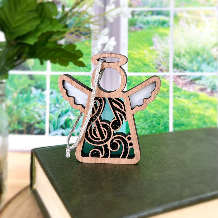 A wooden angel ornament with intricate black and green musical-themed cutouts is displayed on a closed book in front of a window. The background shows a bright, green garden with various plants and a vase with flowers on the left side. This Music Ornament | 3.5" Angel Figurine from the Mother's Angels® collection by Forged Flare® makes an ideal music teacher gift.