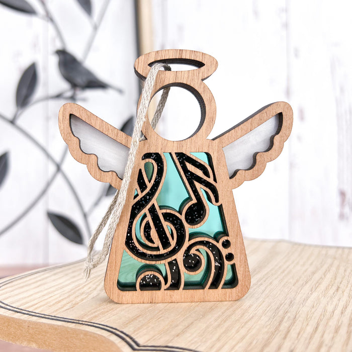 The Music Ornament, a 3.5" Angel Figurine from the Mother's Angels® collection by Forged Flare®, is crafted from wood, featuring cut-out wings and a halo. Adorned with a musical note design at its center, the notes are black with glittery accents against a teal background, and it hangs gracefully by a string. Ideal as a music teacher gift, the backdrop includes an artfully blurred image of a bird perched on a metal branch.