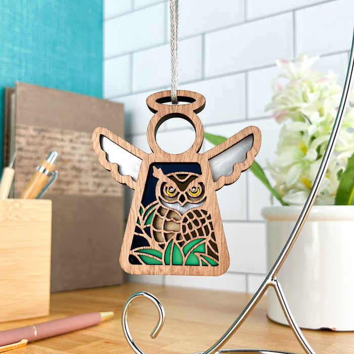 A 3.5" angel figurine from Mother's Angels® by Forged Flare®, this wooden ornament features detailed carvings of an owl in the center and hangs on a string. Perfect as a memorial gift or for bird lovers, it has white wings and green accents on the body. The background showcases a kitchen countertop with a pen, notepad holder, and white tiled wall.