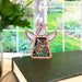 A Forged Flare® Mother's Angels® 3.5" Owl Ornament Angel Figurine, featuring intricate owl carvings and delicate white wings, is propped up on a green book. In the background, a bright window reveals a lush green garden. A glass vase with green leaves and white flowers is partially visible on the left side—a perfect gift for bird lovers or as a memorial tribute.