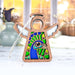 A Peacock Ornament from Forged Flare's® Mother's Angels® collection, featuring a 3.5" angel figurine with an intricate peacock design in green and blue stained glass–style acrylic, stands on a light-colored table. The background showcases a blurry view of a vase and basket, with sunlight streaming through the window in the distance—an ideal gift for any bird lover on any occasion.