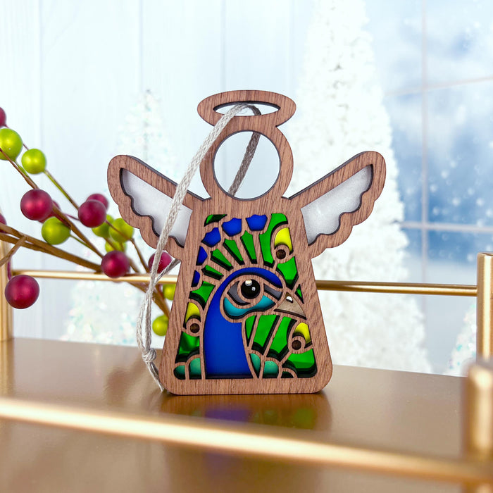 A Peacock Ornament from Forged Flare's® Mother's Angels® collection is elegantly displayed on a brass shelf. This 3.5" angel figurine features white wings and a vibrant stained glass–style body with colorful green, blue, and yellow feathers. In the background, you can see a festive arrangement with red and green berries and snowy trees—a perfect gift for bird lovers.