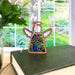 A vibrant Peacock Ornament from the Mother's Angels® collection by Forged Flare®, featuring a 3.5" angel figurine with wooden edges, rests on a closed green book. The backdrop displays a brilliant garden scene through a large window, showcasing lush green foliage and white flowers—ideal as a memorial gift.