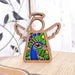 The Peacock Ornament | 3.5" Angel Figurine from Forged Flare®'s Mother's Angels® collection features a wooden angel with cut-out wings, showcasing vibrant stained glass–style acrylic depicting a peacock. Ideal as a memorial gift for bird lovers, the ornament displays white wings and rests on a light wood surface. It is beautifully set against a decorative branch with a bird figure and a rustic white wall in the background.