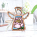 A 3.5" angel figurine from the Mother's Angels® collection by Forged Flare® stands on a white surface. This wooden ornament features wings and a halo, with its middle part showcasing a stained glass–style design of a pink flower with green leaves, reminiscent of the Pink Peony Ornament pattern. The blurred background reveals a potted plant and various objects on a shelf.