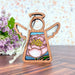 A handcrafted Pink Peony Ornament from Forged Flare®'s Mother's Angels® collection, featuring a 3.5" angel figurine with white wings and a halo, and a stained glass-style depiction of a pink flower on the body, is hanging on a table with a bouquet of purple flowers in the blurred background.