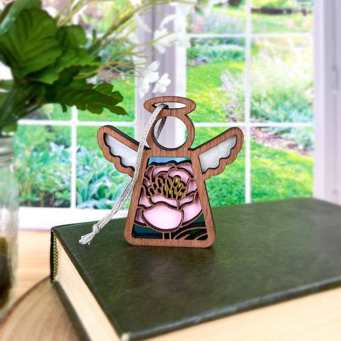 A small 3.5" Mother's Angels® figurine by Forged Flare® stands on a closed book, featuring white wings and a detailed stained glass–style pink peony ornament on its body. Through a large window, the background reveals a vibrant garden scene.