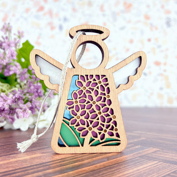 Elevate your festive décor with the Purple Lilac Ornament, a 3.5" angel figurine from the Mother's Angels® collection by Forged Flare®. This wooden ornament boasts intricate floral cutouts and features vibrant shades of purple, green, and blue. It is completed with a braided linen string for easy hanging, while a slightly blurred bouquet of purple flowers adds an extra touch of elegance in the background.