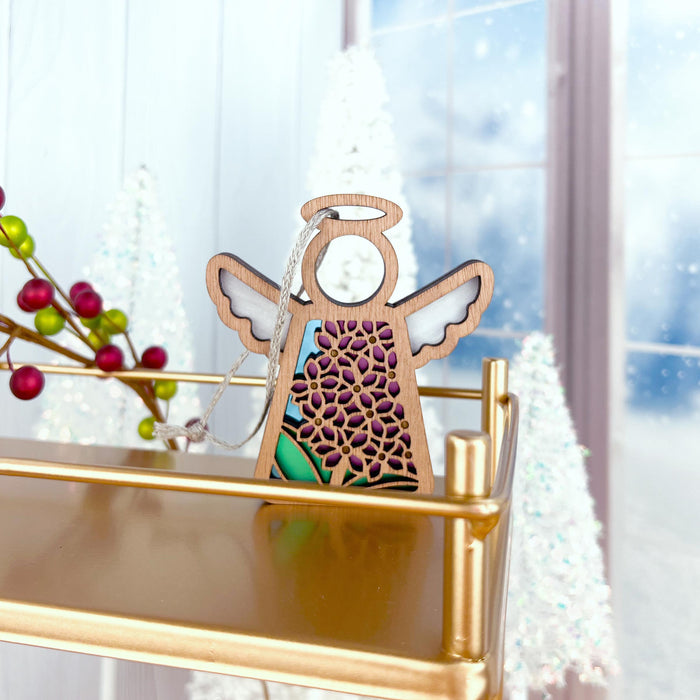 A small, wooden Mother's Angels® Purple Lilac Ornament at 3.5 inches, from Forged Flare®, with floral and colorful accents is hanging on a gold shelf. In the background, white Christmas trees and red berries enhance the festive atmosphere, with a snowy window scene behind them. The setting appears wintery, complemented by delicate angel figurines scattered around.