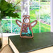 A Forged Flare® Mother's Angels® 3.5" Angel figurine with a wooden frame, adorned with purple flowers and green leaves, stands gracefully on a closed book. The backdrop features a Purple Orchid Ornament amidst floral decor, revealing a window view of a lush garden in full bloom. The scene exudes a peaceful, natural ambiance.