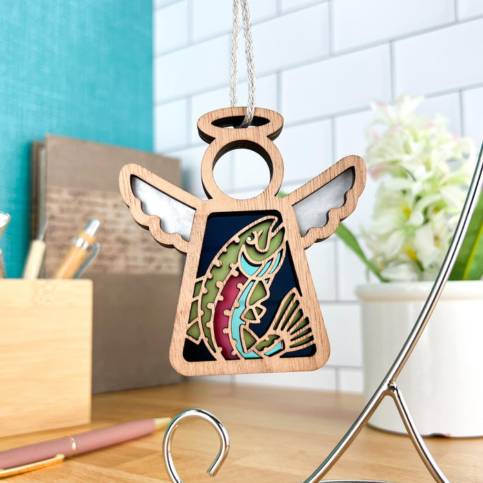 A Rainbow Trout Fish Ornament from Forged Flare®, featuring a vibrant design in the center, hangs in front of a cozy and modern kitchen backdrop. The 3.5" angel figurine, part of the Mother's Angels® collection, has delicate white wings and is suspended by a braided linen string. The contemporary kitchen showcases a white subway tile backsplash and various decor items.