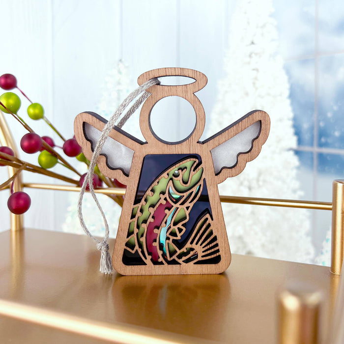 A 3.5" angel figurine from Forged Flare's Mother's Angels® collection, designed with intricate cutouts including a rainbow trout fish ornament, hangs on a stand. The background features blurred white Christmas trees, festive berries on a stick, and a partially visible window showcasing a snowy scene outside.