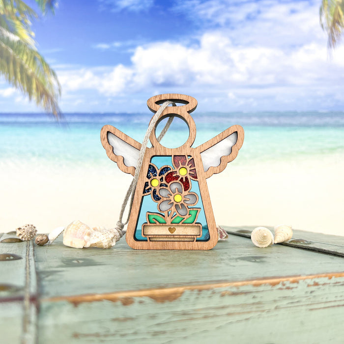 On a beachside table adorned with scattered seashells, stands the Red, White & Bloom Ornament 3.5" Angel Figurine from Forged Flare®'s Mother's Angels® collection. Featuring colorful floral decorations, this charming wooden angel is set against a serene backdrop of turquoise waters, white sand, and palm trees beneath a clear blue sky. This unique piece makes for perfect Patriotic Gifts suitable for any occasion.
