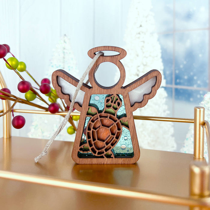 A Sea Turtle Ornament from Forged Flare's® Mother's Angels® collection, featuring a 3.5" wooden angel figurine with white wings and an intricate sea turtle design at its center, is displayed on a wooden surface. A string for hanging is attached to the top. The background showcases bright holiday decor, including red berries and snowy white decorations, making it perfect for beach house decor.