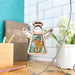 A Seahorse Ornament 3.5" Angel Figurine from Forged Flare® Mother's Angels® collection is hanging on a stand, its turquoise and amber hues sparkling. The backdrop features white tiles, a potted plant, and a desk with pens and a wooden letter holder, making it an elegant display.