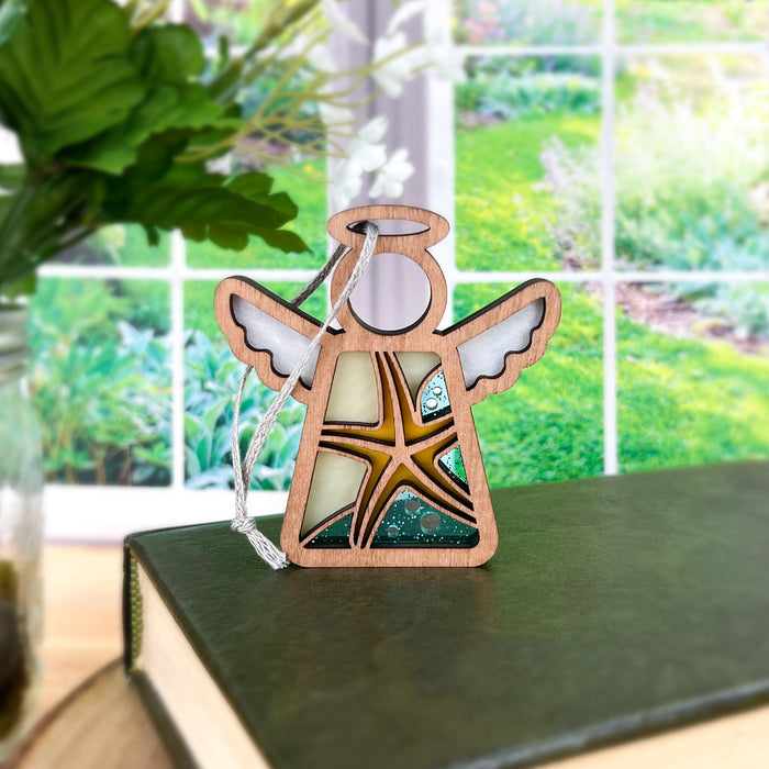 A Starfish Ornament featuring a 3.5" Angel Figurine from the Mother's Angels® collection by Forged Flare® stands gracefully on a closed book, showcasing its wooden craftsmanship and stained glass–style wings with a starfish design in the center. The scene is beautifully complemented by a lush green garden visible through a large window in the background, offering a serene outdoor touch that makes it an ideal choice for those looking for unique Christmas tree ornaments.