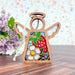 The Strawberry Ornament from Forged Flare's® Mother's Angels® collection features a 3.5" wooden angel figurine adorned with vibrant stained glass–style elements. The angel's body showcases a red strawberry, green leaves, a white flower with a yellow center, and blue background accents, all set against purple flowers and a light speckled wall.