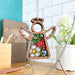 The Strawberry Ornament, a 3.5" angel figurine from Mother's Angels® by Forged Flare®, features decorative stained glass–style wings and dress adorned with red strawberries and a white flower. It hangs elegantly from a string on a metal stand, set against a backdrop of a tiled wall, potted flower, and stationery items on a wooden surface.