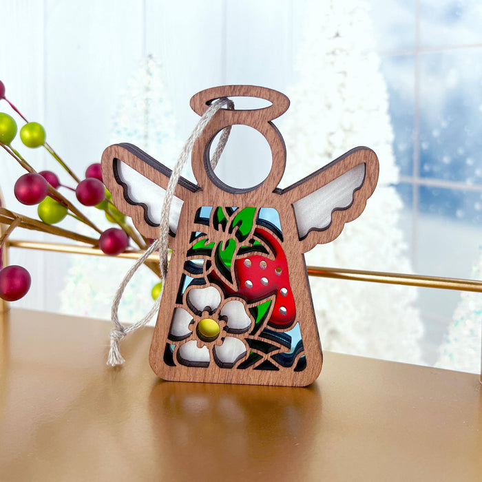 The Strawberry Ornament from Mother's Angels® by Forged Flare® is a 3.5" wooden angel figurine with a stained glass–style effect, showcasing a strawberry, white flower, and green leaves. The angel features white wings and a halo, accompanied by a string for hanging. The background displays blurry, frosted windows and several colorful holiday decorations.