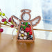 The Strawberry Ornament from Mother's Angels® by Forged Flare® is a 3.5" wooden angel figurine with a stained glass–style effect, showcasing a strawberry, white flower, and green leaves. The angel features white wings and a halo, accompanied by a string for hanging. The background displays blurry, frosted windows and several colorful holiday decorations.