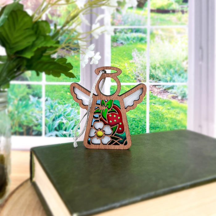 A small 3.5" angel-shaped ornament from the Mother's Angels® collection by Forged Flare®, featuring a colorful strawberry and white flower, stands on a green book near a leafy plant. Set against a blurred background of a bright garden seen through a window, this Strawberry Ornament is perfect for strawberry decor or as unique gift ideas for women.