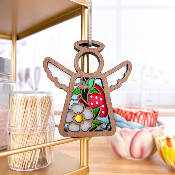 A charming 3.5" angel figurine from the Mother's Angels® collection by Forged Flare®, this decorative Strawberry Ornament features a delightful depiction of a strawberry and a flower. Hanging on a string, it is elegantly displayed on a golden shelf in a bright kitchen setting with bowls and jars in the background, making it an ideal Christmas tree ornament or thoughtful strawberry gift.