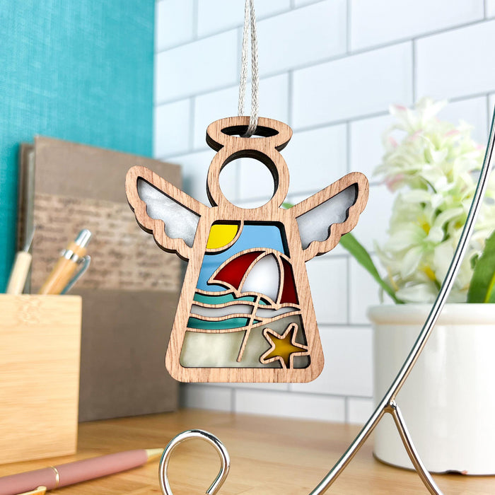 The Summer Beach Ornament | 3.5" Angel Figurine by Mother's Angels® from Forged Flare® is a charming wooden angel decoration that depicts a serene beach scene complete with a sun, umbrella, waves, and starfish. Hangs elegantly on a string, it's placed on a desk adorned with books, pens, and a potted plant against a tiled wall. It serves perfectly as both beach house decor and Christmas ornament.