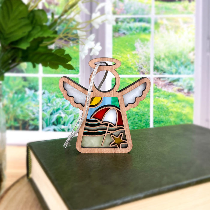 A Forged Flare® Summer Beach Ornament, a 3.5-inch angel figurine from the Mother's Angels® collection, stands on a closed book. This wooden angel features a sun, sailboat, and starfish. The background reveals a lush garden through a window, with vibrant green plants and flowers. Perfect as beach house decor or to adorn your Christmas tree.