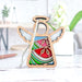 The Summer Watermelon Ornament by Forged Flare® is a 3.5" angel figurine from the Mother's Angels® collection. This charming wooden Christmas tree ornament features vibrant stained glass–style wings and body, with segments in red, green, and blue. It includes a braided linen string at the top for easy hanging. The background setting is a softly blurred white room with a plant and a pen on a table.