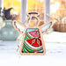 A Summer Watermelon Ornament by Forged Flare®, featuring a 3.5" angel figurine from the Mother's Angels® collection, sits on a white surface. The background displays blurred autumnal colors seen through a window, complemented by decorative items such as vases. This charming Christmas tree ornament has a rustic and festive appearance with its stained glass–style design in red, green, and white.