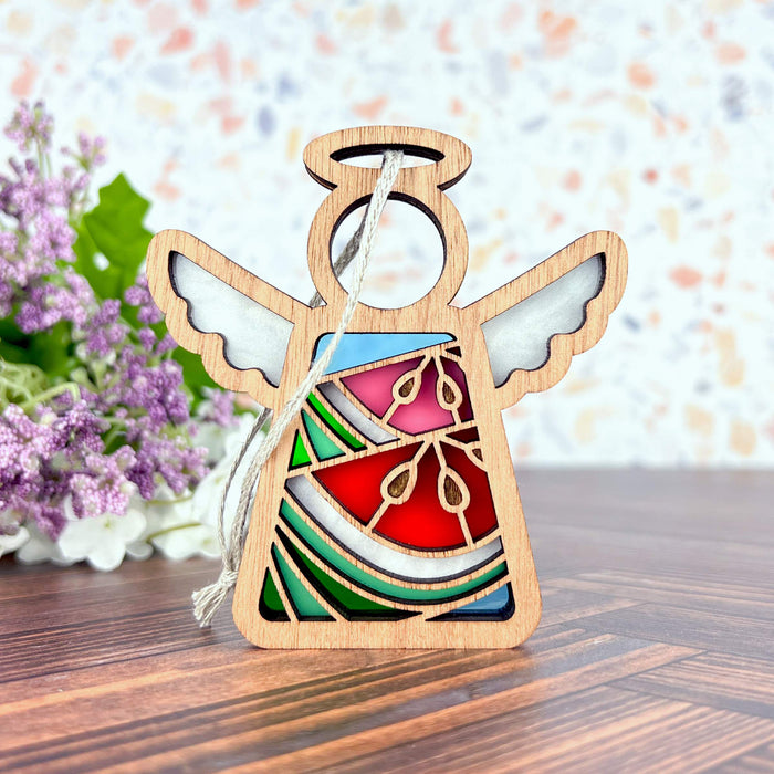 A Summer Watermelon Ornament, a 3.5" angel figurine from the Mother's Angels® collection by Forged Flare®, boasts stained glass–style wings and gown with vibrant sections of red, green, and blue. It hangs elegantly from a string against a terrazzo pattern wall backdrop and is complemented by a bouquet of purple flowers on a wooden surface—making it the perfect addition to your Christmas tree for adding festive charm.