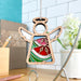 The Summer Watermelon Ornament by Forged Flare® is a 3.5" angel figurine with stained glass–style wings and a colorful, geometric dress that hangs from a string. In the background, you can see a modern kitchen with white tiles, a potted plant, and various stationery items on a wooden countertop—making it an ideal Christmas tree ornament.