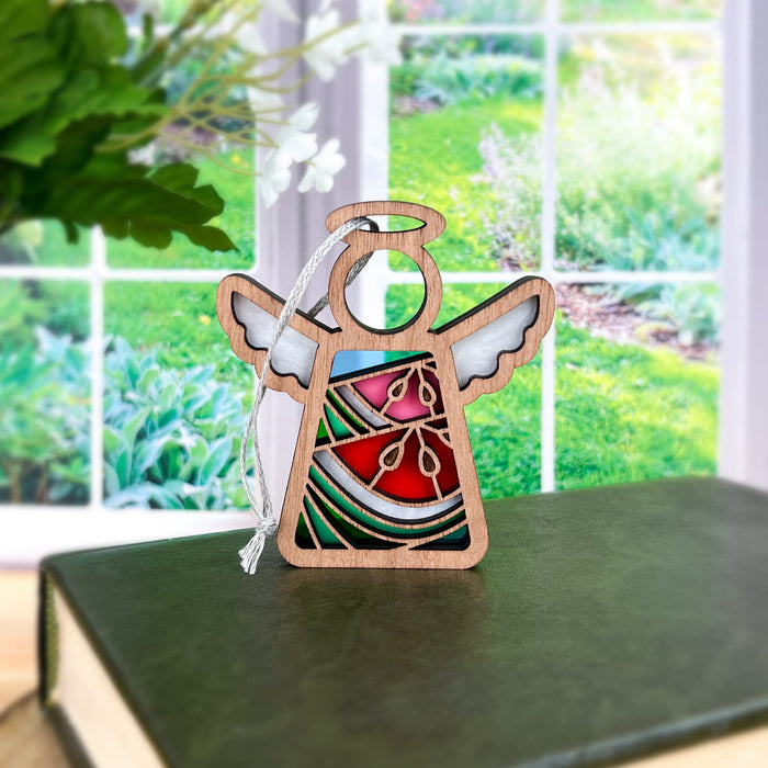 A Summer Watermelon Ornament from Forged Flare®, a 3.5" angel figurine from the Mother's Angels® collection, featuring vibrant stained glass–style designs, is placed on a green book in front of a window. Lush greenery and plants can be seen through the window, creating a serene outdoor backdrop.