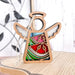 The Summer Watermelon Ornament | 3.5" Angel Figurine from the Mother's Angels® collection by Forged Flare® is a beautiful wooden angel ornament with stained glass–style wings and body in vibrant red, green, and blue colors, adorned with a golden branch design. This festive Christmas tree ornament stands elegantly on a wooden surface, complemented by a decorative bird wall hanging in the background.