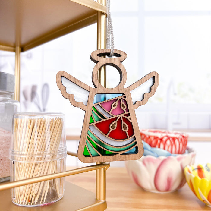 A Summer Watermelon Ornament from the Mother's Angels® collection by Forged Flare®, featuring a 3.5" angel figurine, gracefully hangs on a gold-colored shelf in a bright kitchen. Nearby, a container of toothpicks, a glass jar, and patterned bowls can be seen, adding a cozy and decorative touch to the space, reminiscent of charming Christmas tree ornaments.