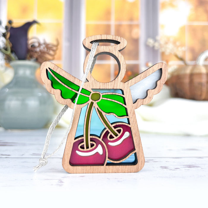 The Sweet Cherries Ornament from Mother's Angels® by Forged Flare® features a 3.5" wooden angel figurine adorned with colorful stained glass–style accents and sweet cherries with green leaves. Hanging by a string, the ornament's blurred indoor background showcases vases and autumnal decor, making it an enchanting addition to any Christmas tree.