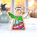 The Sweet Cherries Ornament from Mother's Angels® by Forged Flare® features a 3.5" wooden angel figurine adorned with colorful stained glass–style accents and sweet cherries with green leaves. Hanging by a string, the ornament's blurred indoor background showcases vases and autumnal decor, making it an enchanting addition to any Christmas tree.