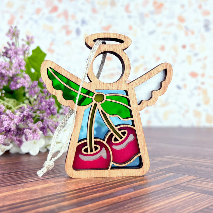 A Sweet Cherries Ornament by Forged Flare®, featuring a 3.5" Mother's Angels® angel figurine with stained glass–style pieces in green, red, and blue, sits on a wooden surface. The Christmas tree ornament includes a design of cherries. In the background, there's a bouquet of purple flowers with lush green leaves against a softly multicolored wall.