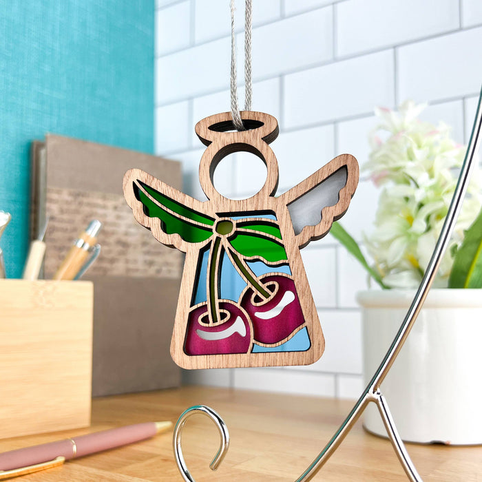 The Sweet Cherries Ornament from the Mother's Angels® collection by Forged Flare® is a 3.5-inch wooden angel figurine, featuring colorful stained glass-like wings and sweet cherries on its body, hanging on a metal stand. It is displayed on a wooden surface with office supplies and a potted white flower in the background.