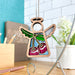 The Sweet Cherries Ornament from the Mother's Angels® collection by Forged Flare® is a 3.5-inch wooden angel figurine, featuring colorful stained glass-like wings and sweet cherries on its body, hanging on a metal stand. It is displayed on a wooden surface with office supplies and a potted white flower in the background.