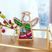 The Sweet Cherries Ornament, a 3.5" wooden angel figurine from Mother's Angels® by Forged Flare®, is displayed on a surface. Its winged design features colorful cherries on its body. In the background, festive red and green berries are placed alongside a snowy windowpane and white Christmas trees outside, creating a holiday ambiance. This ornament makes for meaningful gifts for Christians.