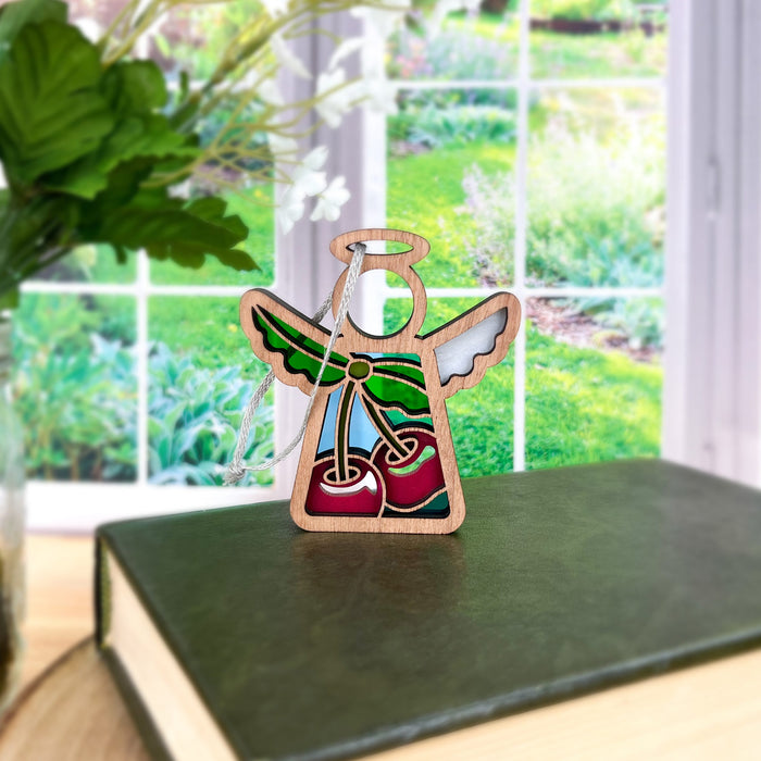 A small, colorful Sweet Cherries Ornament - a 3.5" angel figurine from the Mother's Angels® collection by Forged Flare® - stands on a green hardcover book. Behind the book is a bright window, offering a view of a lush green garden. The background features green leaves and white flowers in a vase on a wooden table, perfect for highlighting angel figurines.