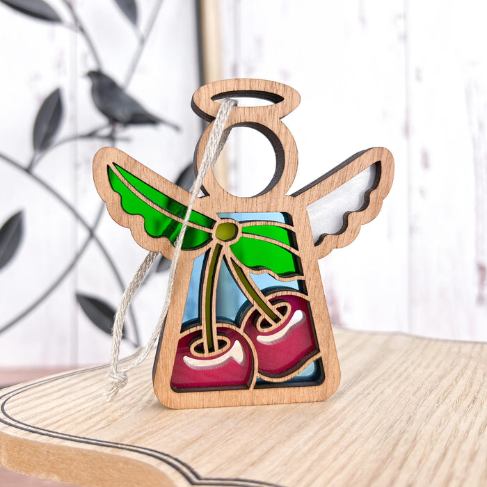 The Forged Flare® Sweet Cherries Ornament | 3.5" Angel Figurine from the Mother's Angels® collection showcases a wooden angel with hollow wings and head, adorned with stained glass–style cherries. The body features a depiction of two charming cherries accompanied by green leaves. Placed on a light-colored wooden surface, the Christmas tree ornament has a blurred bird decoration in the background.