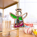 A Sweet Cherries Ornament from the Mother's Angels® collection by Forged Flare® hangs from a shelf in a cozy kitchen. This 3.5" angel figurine, featuring colorful wings and cherries on its dress, is surrounded by various kitchen items, including toothpicks and bowls with flower designs in the background. This charming piece makes an ideal Christmas tree ornament or thoughtful gift for Christians.
