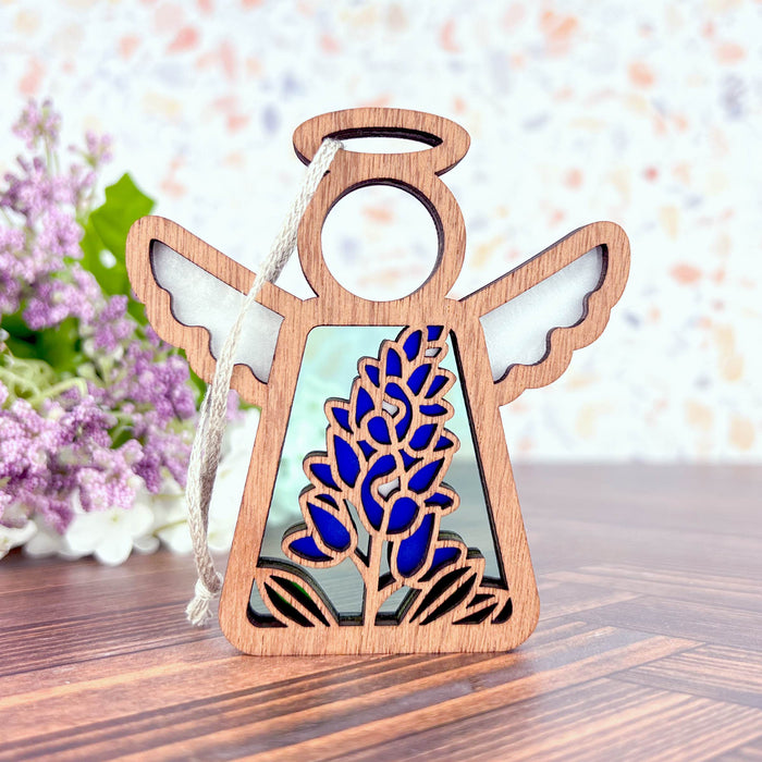 The Texas Bluebonnet Ornament by Forged Flare® is a 3.5-inch wooden angel figurine from the Mother's Angels® collection, featuring cutout wings and a halo. The angel's body showcases a stained glass–style insert depicting a Texas bluebonnet flower. This Christmas tree ornament includes a string for hanging and is set against a backdrop of lavender flowers and a light, speckled wall.